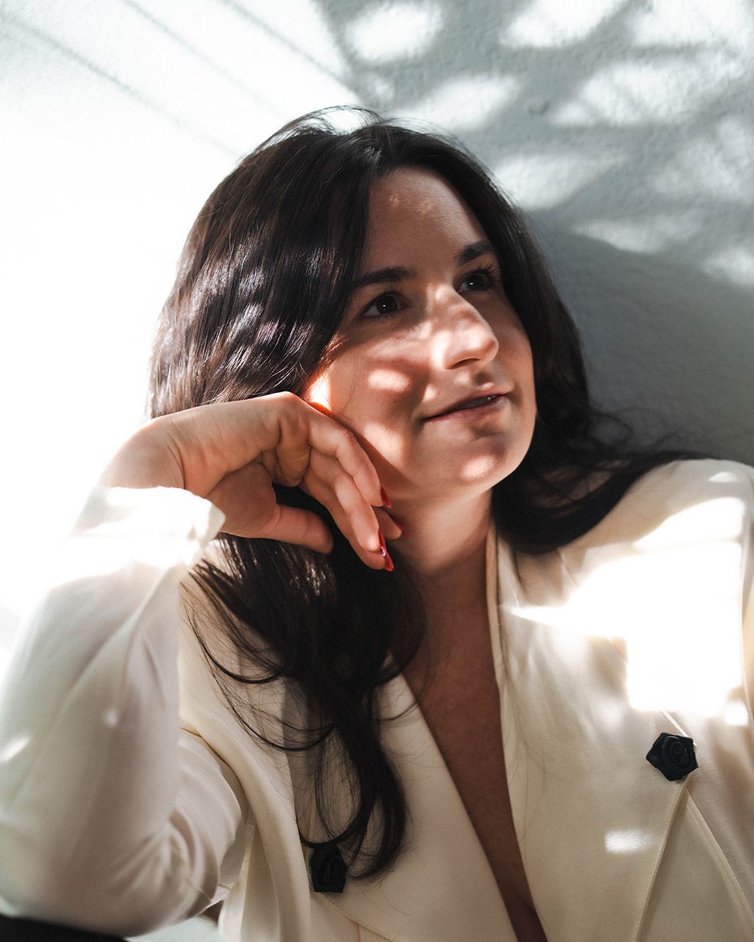 Jade Levinson, somatic intimacy coach, smiling in a white blazer with leaf shadows on the wall