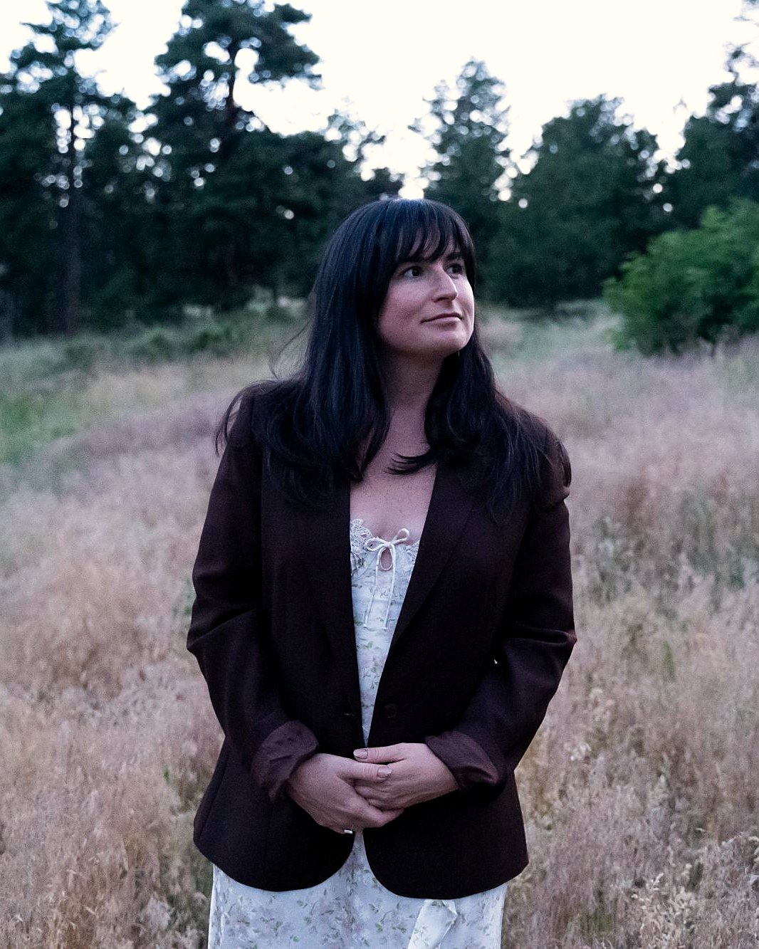 Jade Levinson in a dark blazer standing in a meadow of tall grass at dusk, looking off to the side