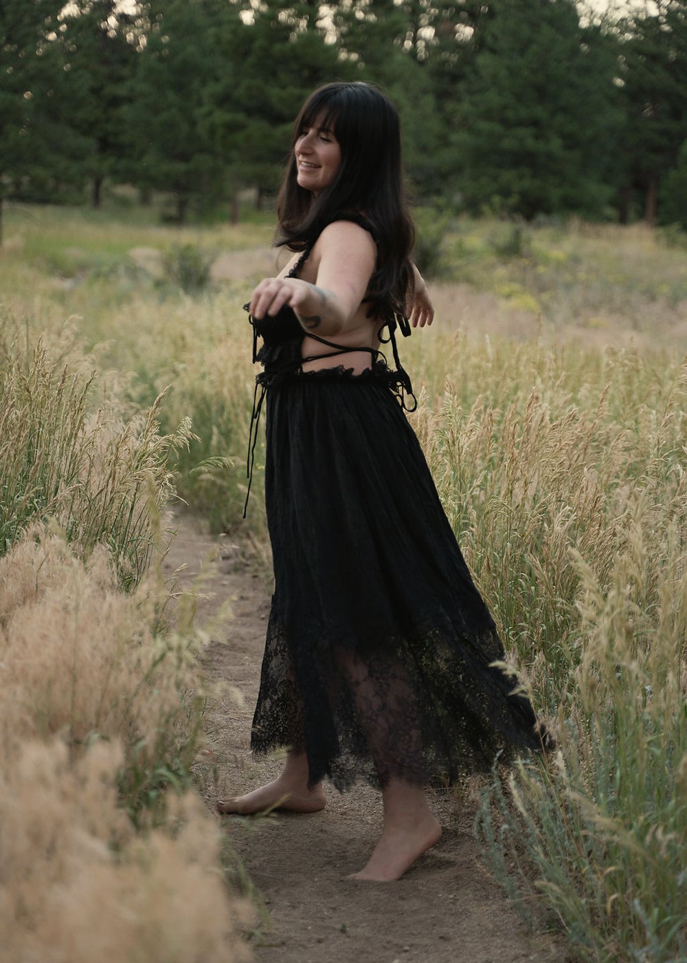 Jade Levinson barefoot on a meadow path in a black lace dress, arms open, smiling