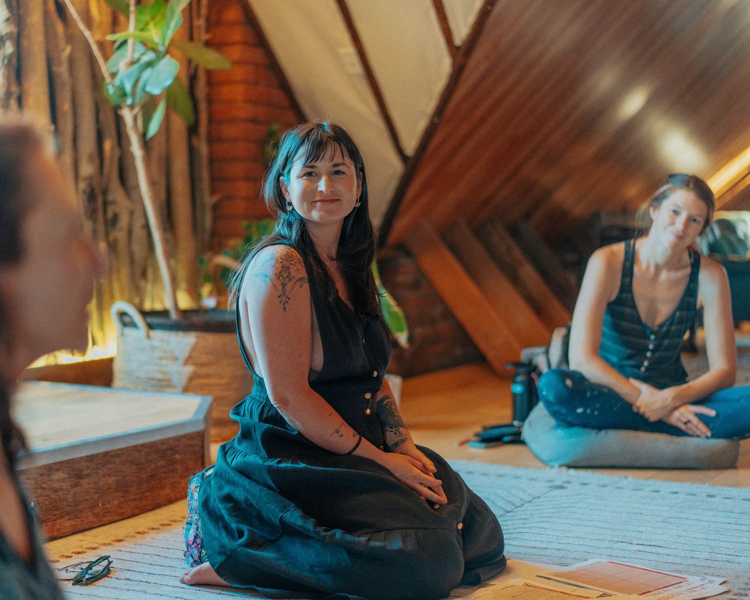 Jade Levinson kneeling on a rug in a warm, plant-filled workshop room, smiling at participants seated around her